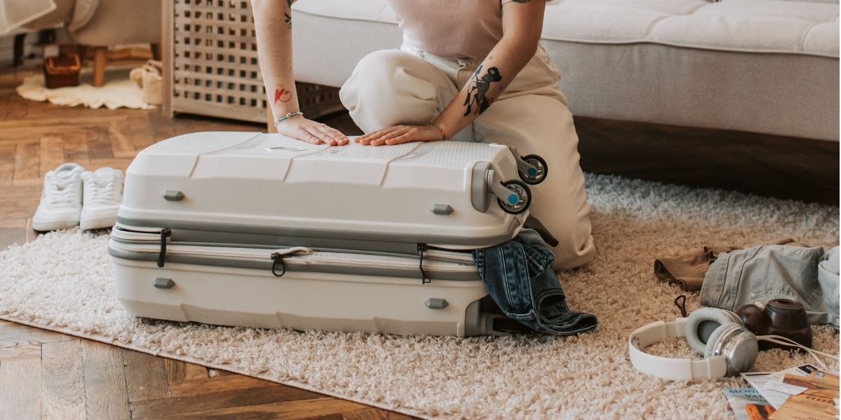 packing suitcase woman trying to close a full suitcase with some clothes spilling out and a pile of other clothing and shoes on the floor near her